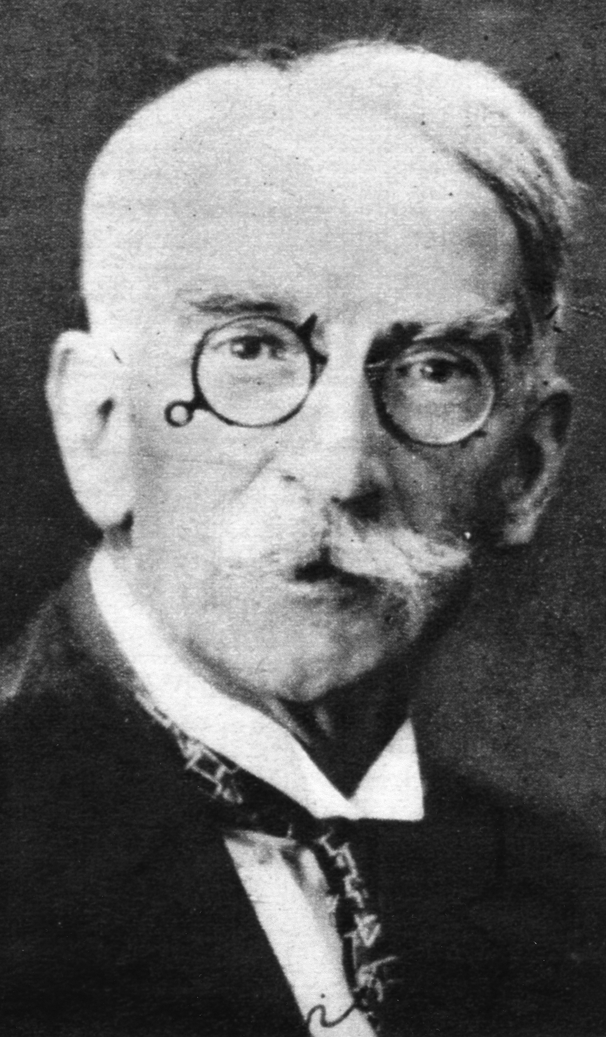 Portrait of an older man with round spectacles wearing an old fashioned suit and tie. He has neat white hair and a bushy white mustache. 