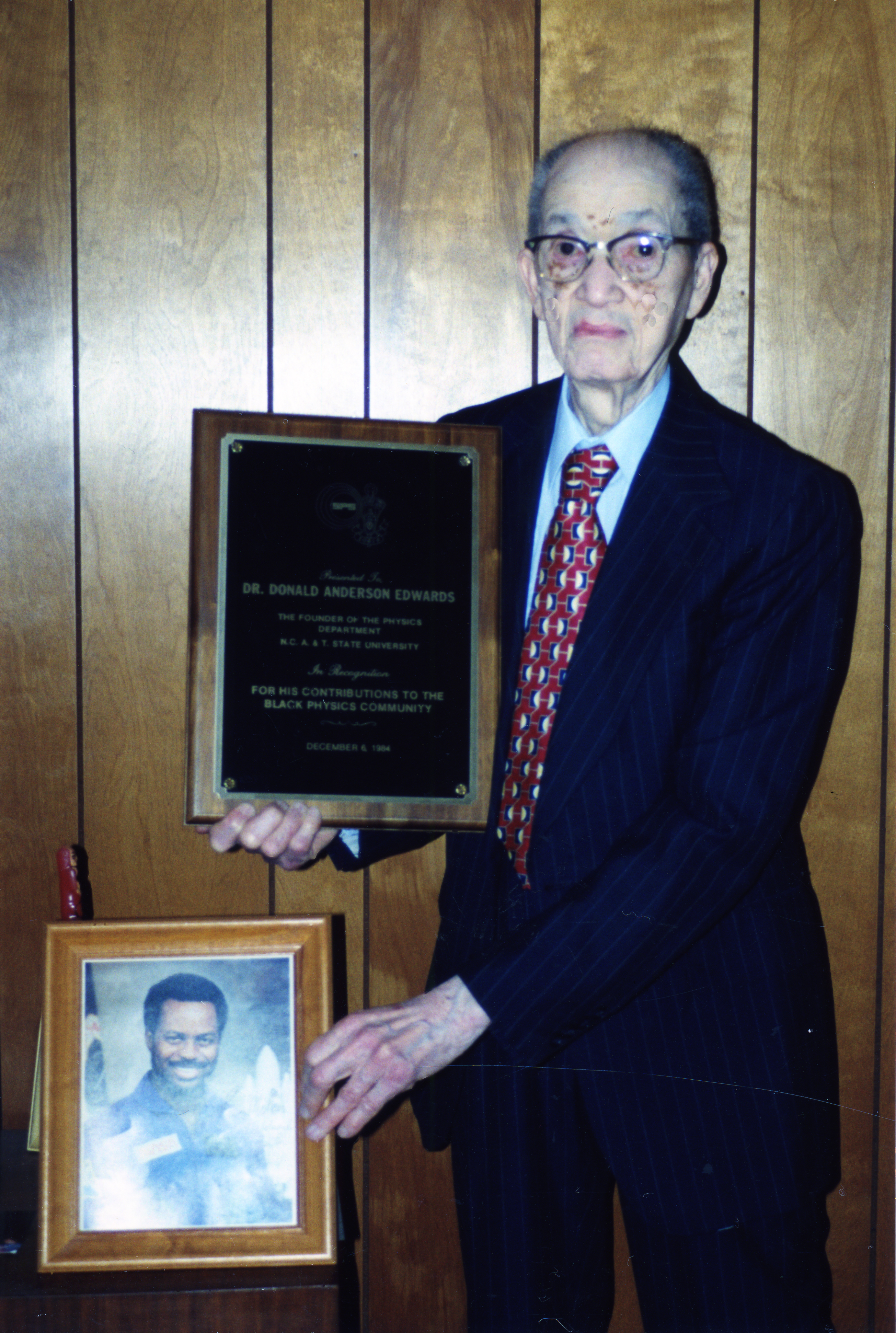Donald A. Edwards stands before a wood paneled wall, holding a plaque and a photo of Ronald McNair.