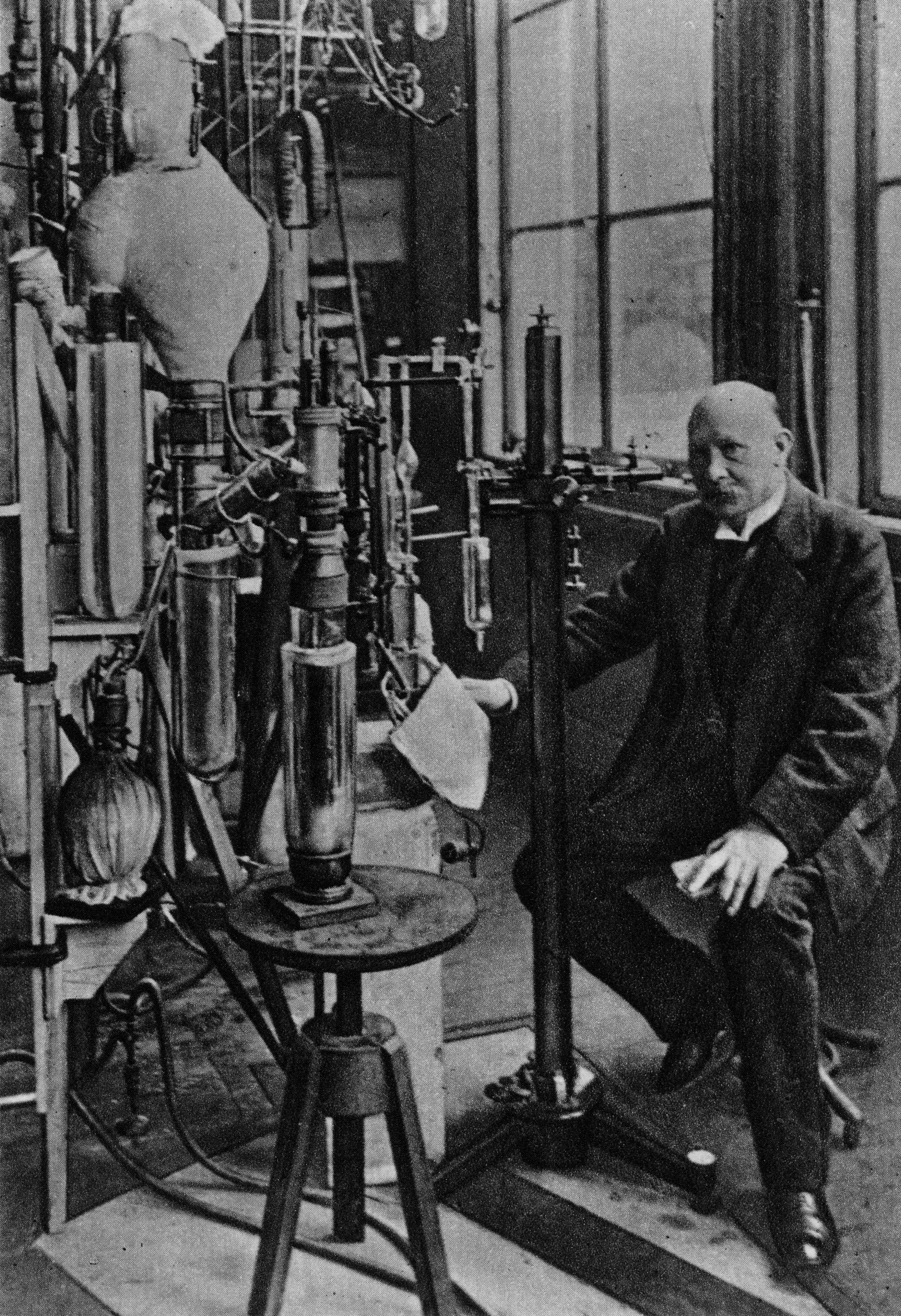 a balding older white man in a dark suit sits on a stool next to a bunch of tube-like equipment