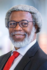 A headshot of Jim Gates, a Black man smiling, wearing a suit, with greying hair.