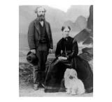 A black and white posed photograph of James Clerk Maxwell standing in a three-piece suit, holding a hat in one hand. Katherine Maxwell sits in a dark dress, a large cross hanging from her neck. At her feet is a very shaggy small white dog. The backdrop is a scene of rocks by the ocean.