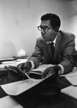 A middle-aged African American man wearing glasses and a suit and tie sits at a desk covered in papers. He gestures at one of the papers with a pencil.