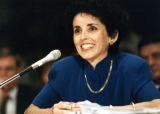 France Córdova, wearing a gold necklace over a smart blue blazer, leans towards a table-top microphone