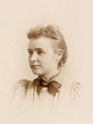 Portrait of a young woman in three-quarter profile wearing a light-colored dress with a dark bow tie circa the late 1800s. Her hair is pulled back except for short curly bangs in the front