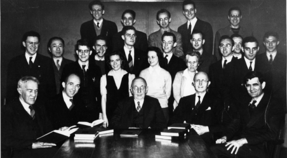 a black and white group photo of 20 men and three women standing and sitting around a table