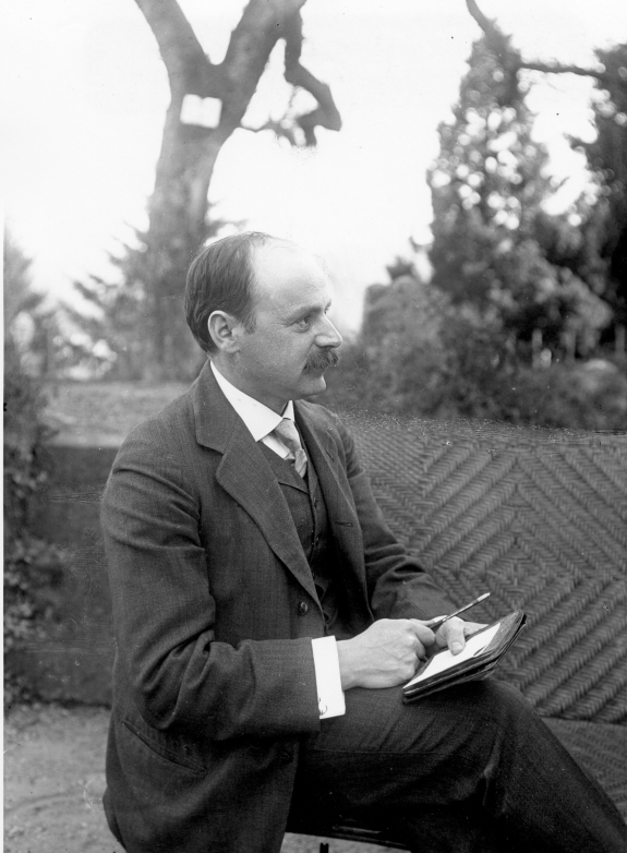 a man with dark hair and wearing a suit sits outdoors holding a pen and a piece of paper with a small notebook, as if he has just written something or is about to write