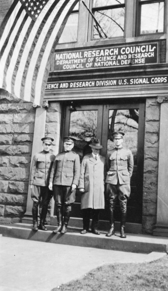 four men stand on the doorstep of a building, three of the men (including Robert Millikan) wear US army uniforms in khaki with high polished boots and caps, the fourth man wears a long civilian coat and hat. above the door way two signs read "National Research Council: Department of Science and Research: Council of National Defense" and "Science and Research Division U.S. Signal Corps." projecting out from the doorway is a flagpole with a large U.S. flag