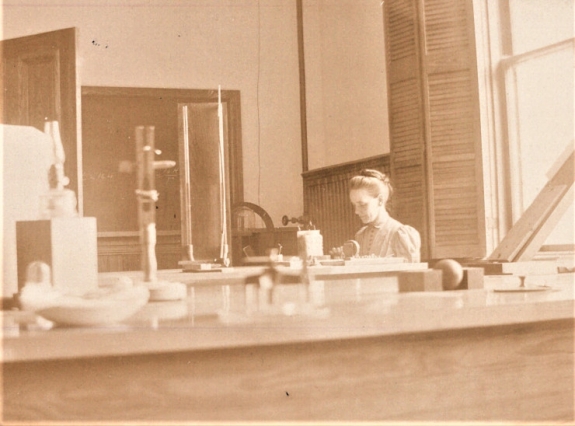 In this sepia photograph, the camera peaks over the top of a lab bench to capture Marcia Keith sits with head bent amidst laboratory equipment at Mount Holyoke College. 