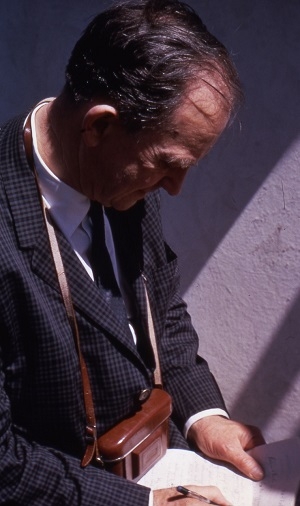 Image of Merle Tuve, with a camera around his neck, writing notes in 1966. AIP Emilio Segrè Visual Archives, John Irwin Slide Collection 