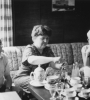 From left to right, Yakov Zeldovich, Alla Genrikhovna Masevich, and Vera Rubin sit on a couch around a coffee table decked with a china tea set. Zeldovich and Rubin look towards the camera smiling while Masevich opens a bottle of a cold drink. 