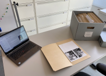 A gray table with, from left to right, a laptop, and open manila folder, and an open carton box with folders inside it. A file cabinet is visible in the background.