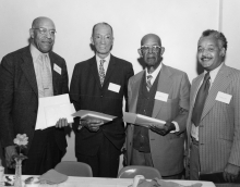 Four men stand side-by-side holding pieces of paper; all are in suits