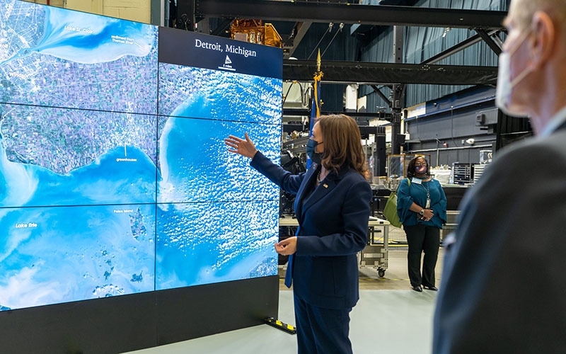 Harris gesturing in front of a screen displaying satellite images with onlookers