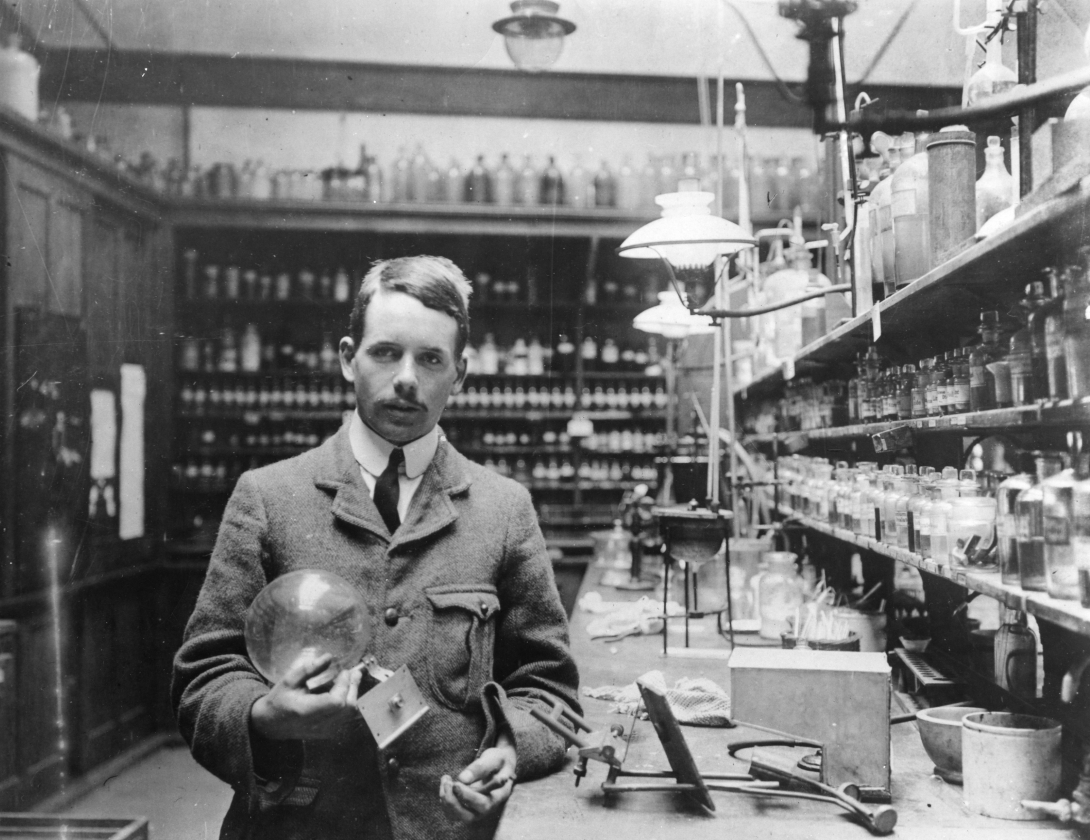 a young man with dark hair and a mustache wearing a practical suit holds x-ray tubes in a laboratory with other kinds of glassware and laboratory equipment crowding the shelves and the laboratory bench he stands next to
