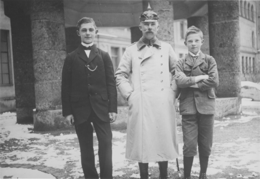 two young boys stand on either side of an older man wearing a german military uniform, pointed helmet, and long winter coat. They stand outside in front of a grand stone building and there is snow on the ground