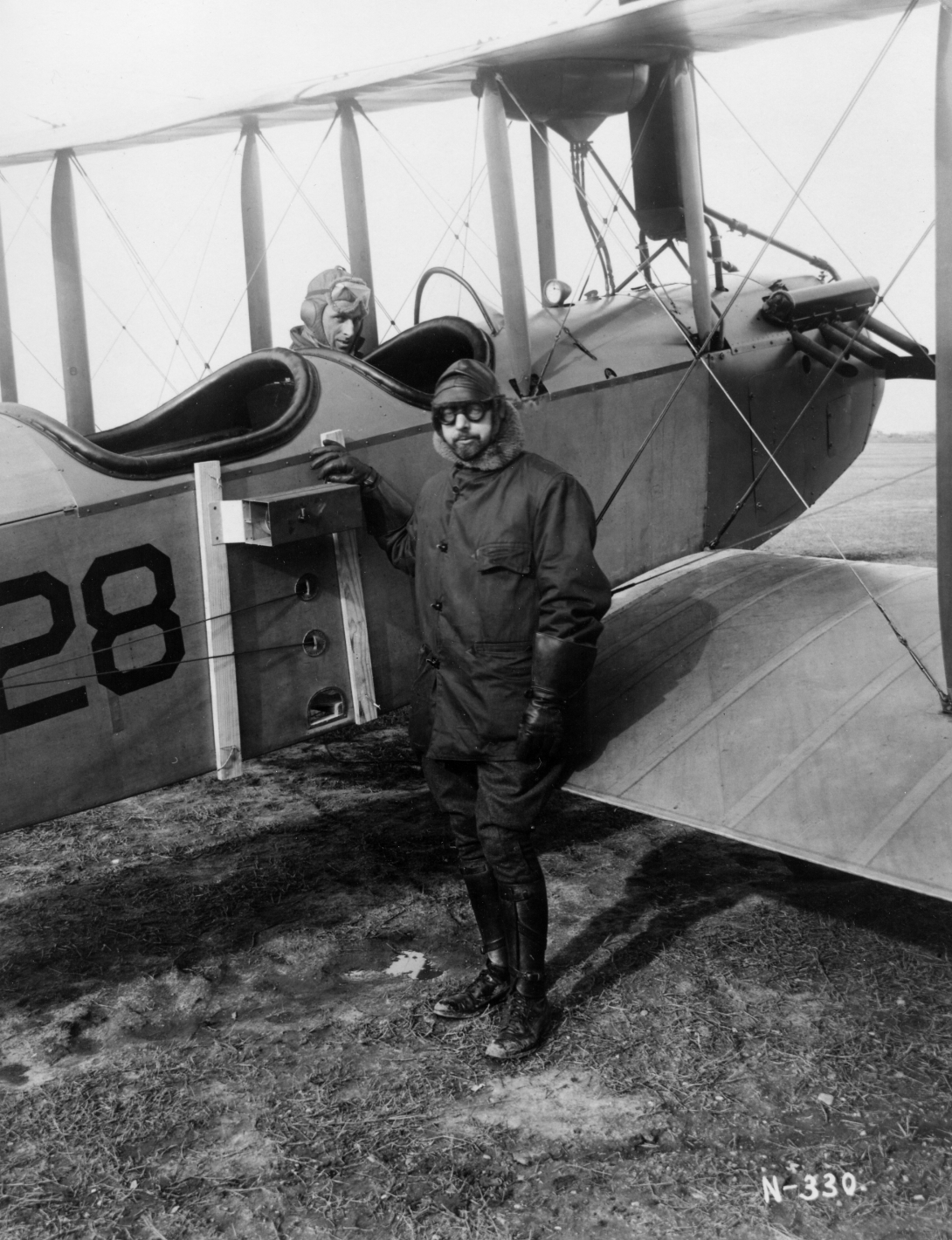 a man wearing thick glasses,flying cap and jacket, along with long gloves and boots, stands next to a camera attached to the side of a two-seater biplane. in the front seat of the plane sits a pilot with aviator cap and goggles