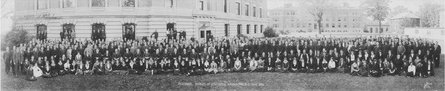 hundreds of individuals in 1920 era clothing sit and stand for a group portrait on a lawn in front of a brick and stone building, with other parts of the lawn and mroe buildings in the background. Perhaps 80 of the 3-400 people in the photo are women