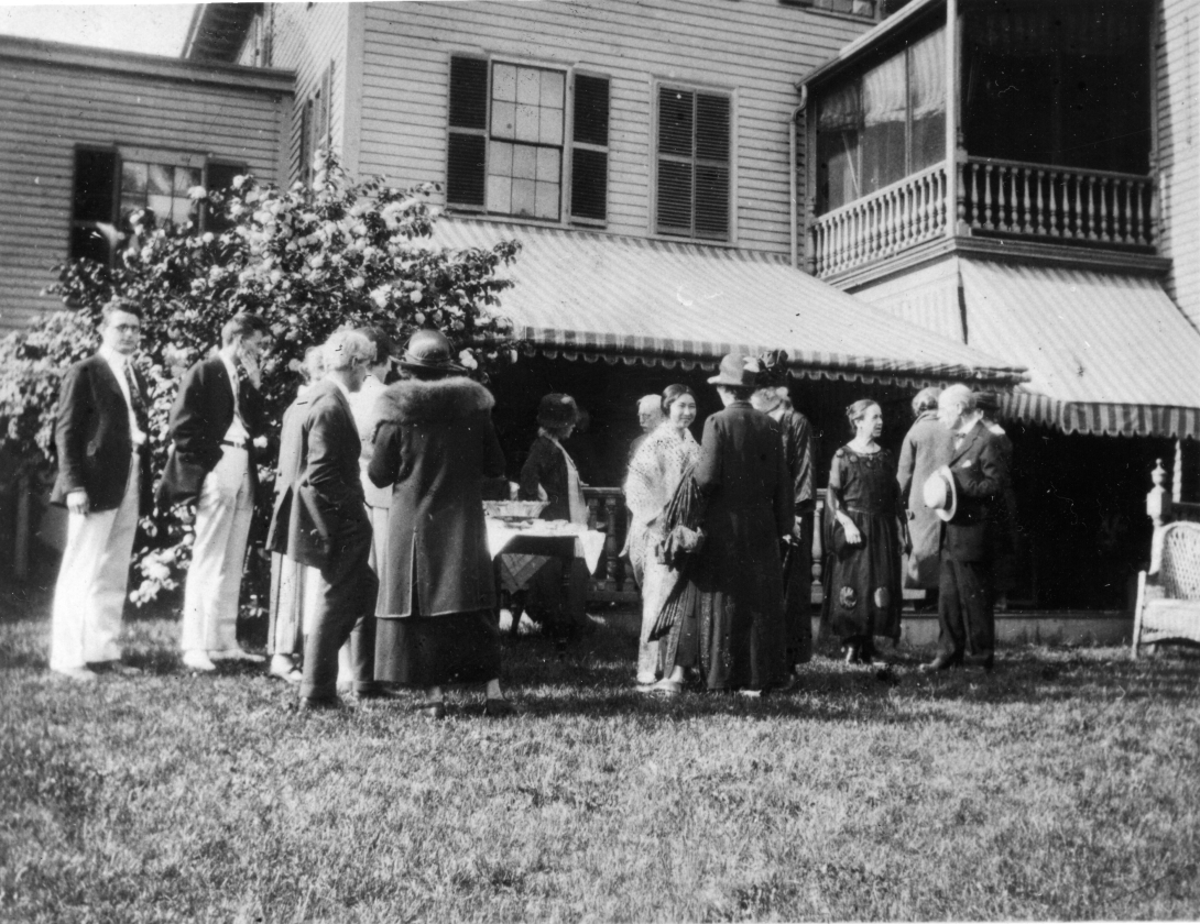 Men in suits and women in dresses stand around chatting on a lawn in front of a house. in the background a table with food and drinks is visible.