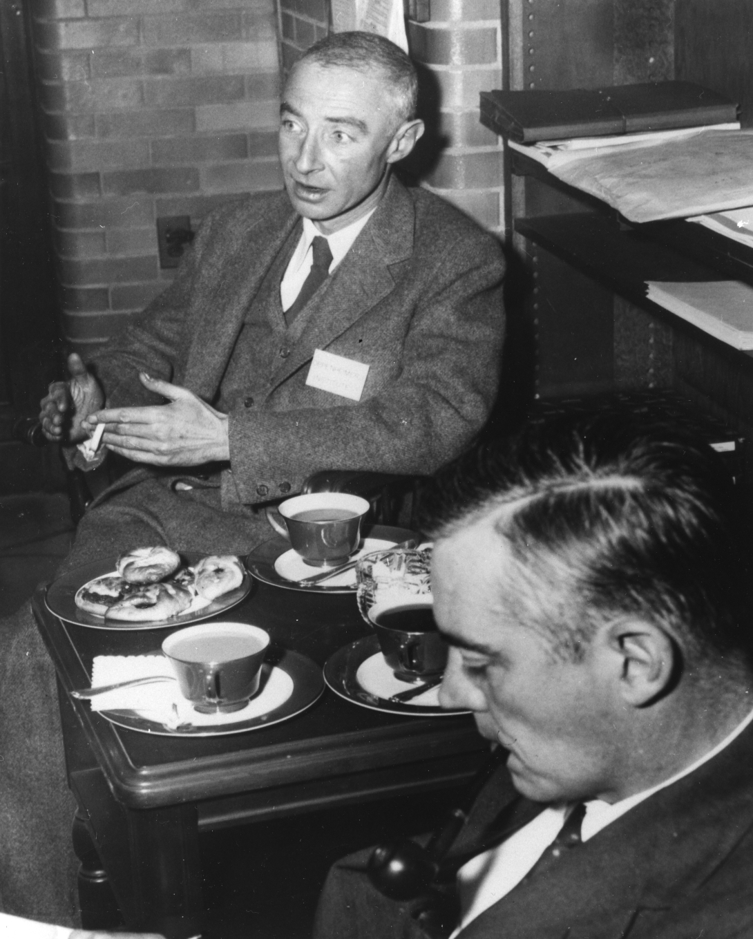 Oppenheimer and another man, both in suits, sit at a small table with cups of tea and plates of pastries in front of them. Oppenheimer wears a nametag and gestures while he talks to someone off camera. The second man is in the foreground looking down while he smokes a pipe.
