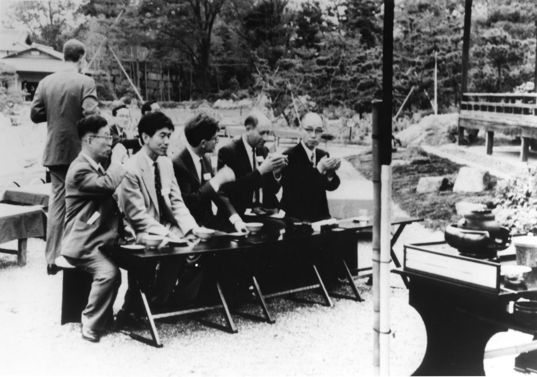 five men in suits and nametags sit on stools in front of a long table. they hold cups of tea in their hands. In the background are decorative paths and plants. A building is just visible in the background through some trees. 