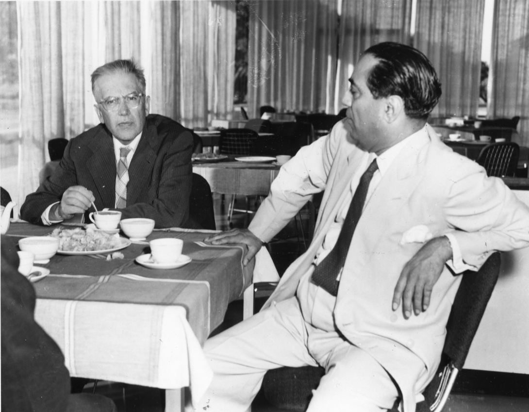 In a cafeteria with floor-to-ceiling windows and curtains, Emilio Segre sits at a table and stirs his cup of tea. To his left sits Homi Bhabha, who leans back in his chair with another cup of tea in front of him. A third man at the table has his back to the camera.