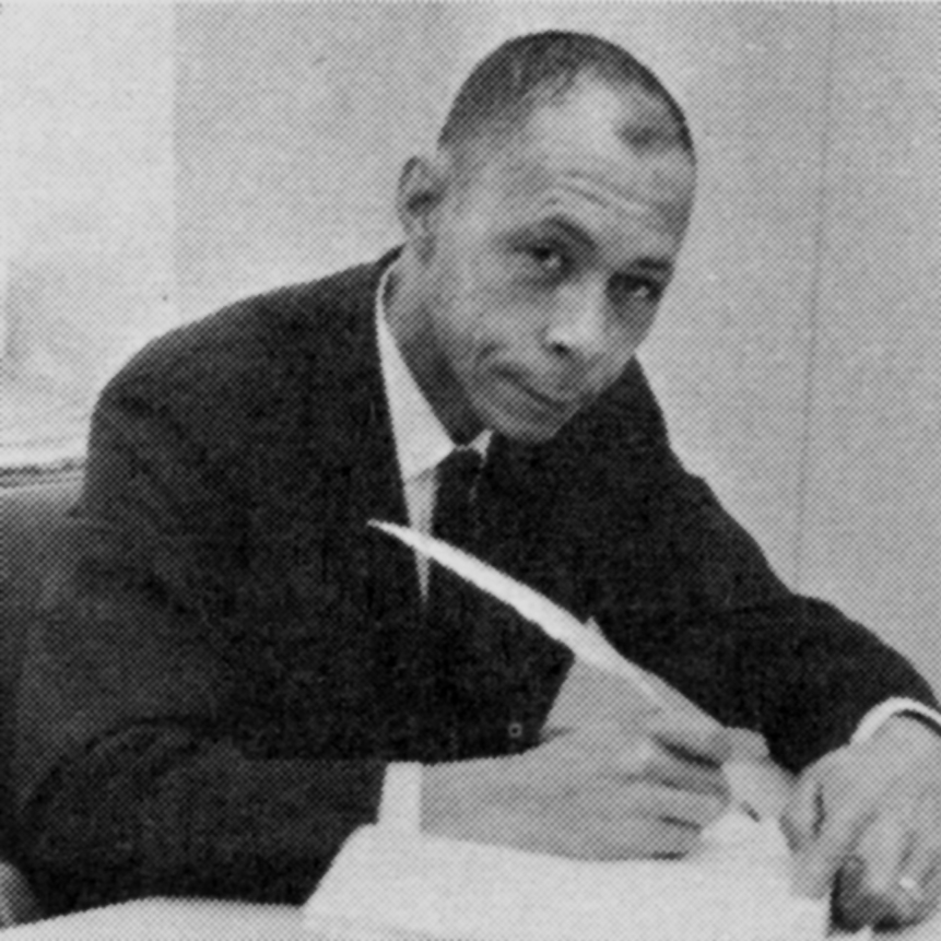 Black-and-white photo of Titus Pankey, Jr., wearing a suit and sitting while holding a feathered quill pen. at a desk. He is holding a 