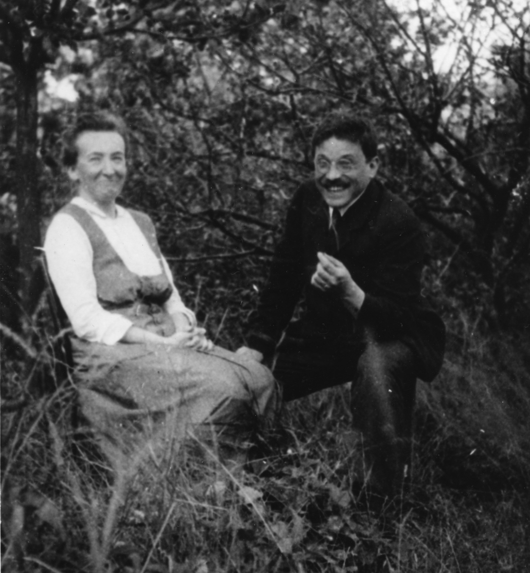 A woman in a dress sits in a garden. She looks mildly amused or trying not to laugh, while next to her kneels a man in a suit with a big bushy mustache laughs openly.