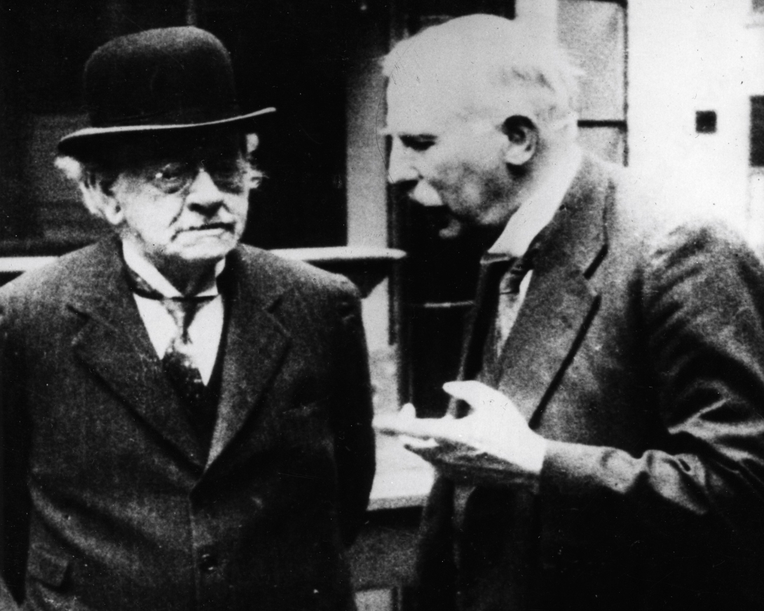 two men in old fashioned suits stand conversing. J.J. Thomson, on the left, wears eyeglasses and a bowler hat and looks pensive. Ernest Rutherford, standing on the right, gestures drawing attention to his bushy white mustache.