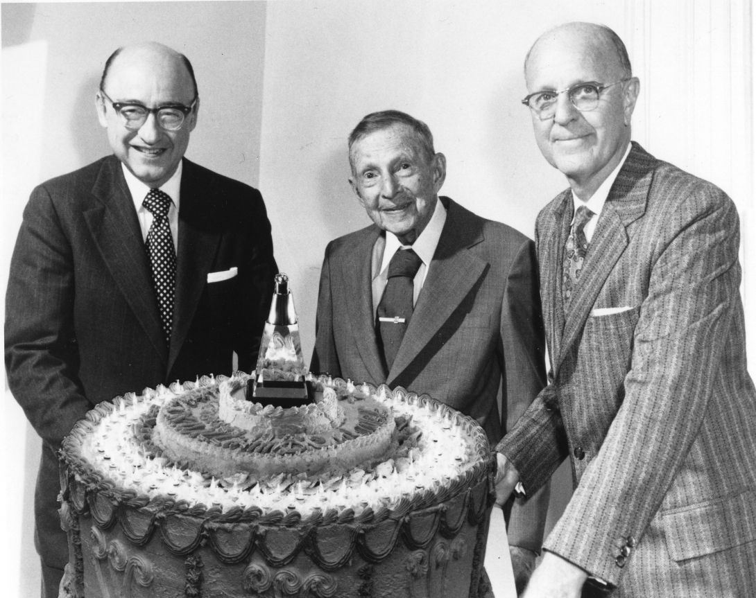 three older men (the one in the center looks very elderly) stand around a massive birthday cake. there are so many lights on the cake that it looks like the whole thing is on fire