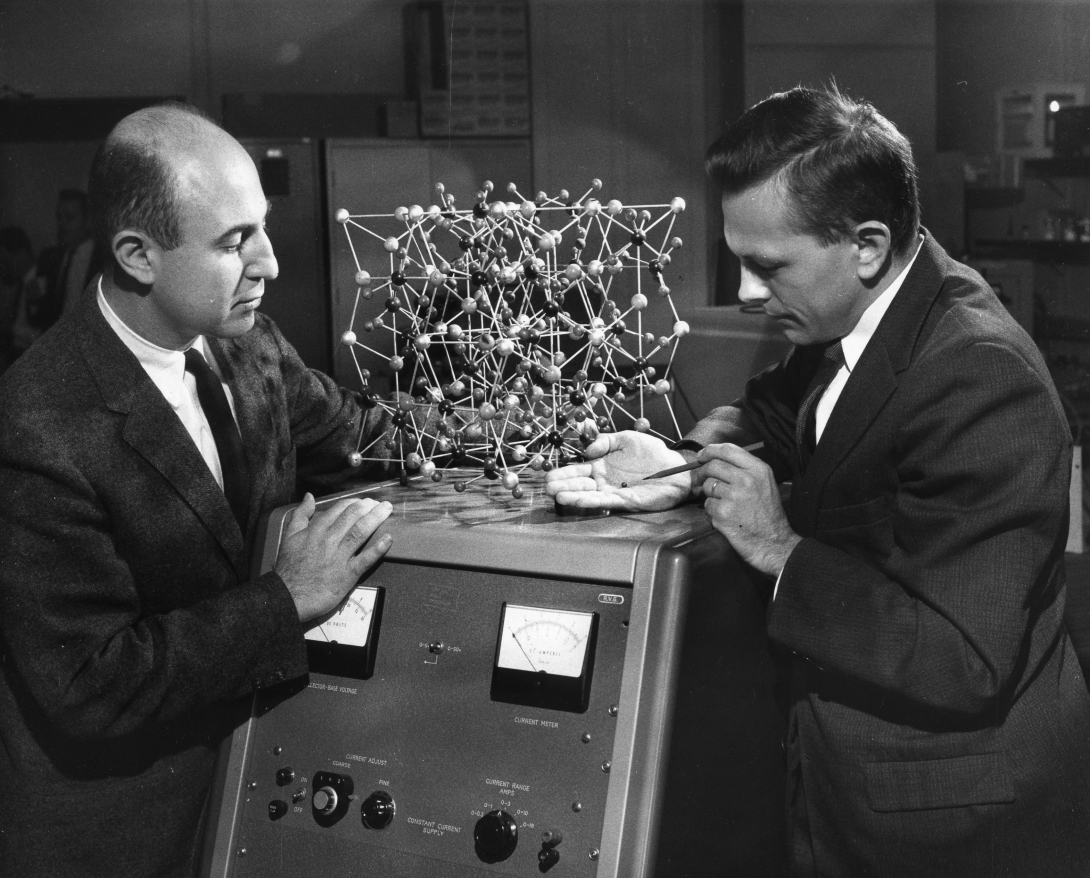 two man in suits stand on either side of a cell model examining it 