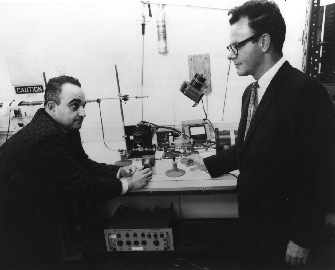 two men in suits, one sitting and one standing, lean next to a laboratory bench with equipment on it