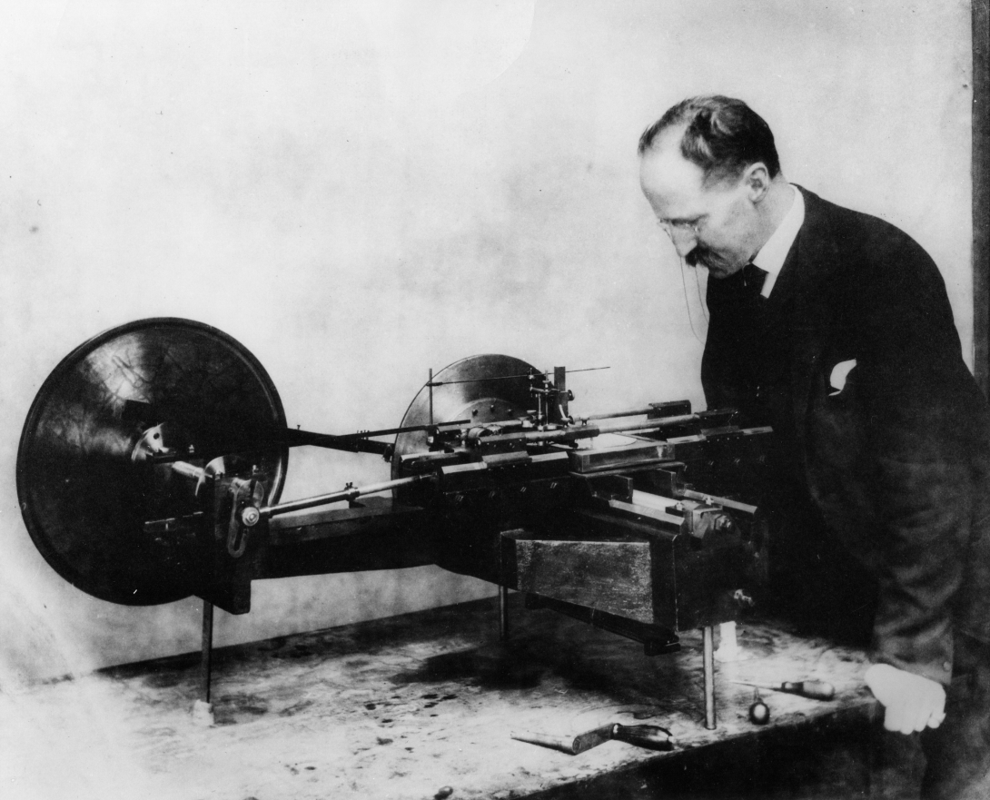 a man in a late 1800s suit leans over a piece of equipment with big wheels 