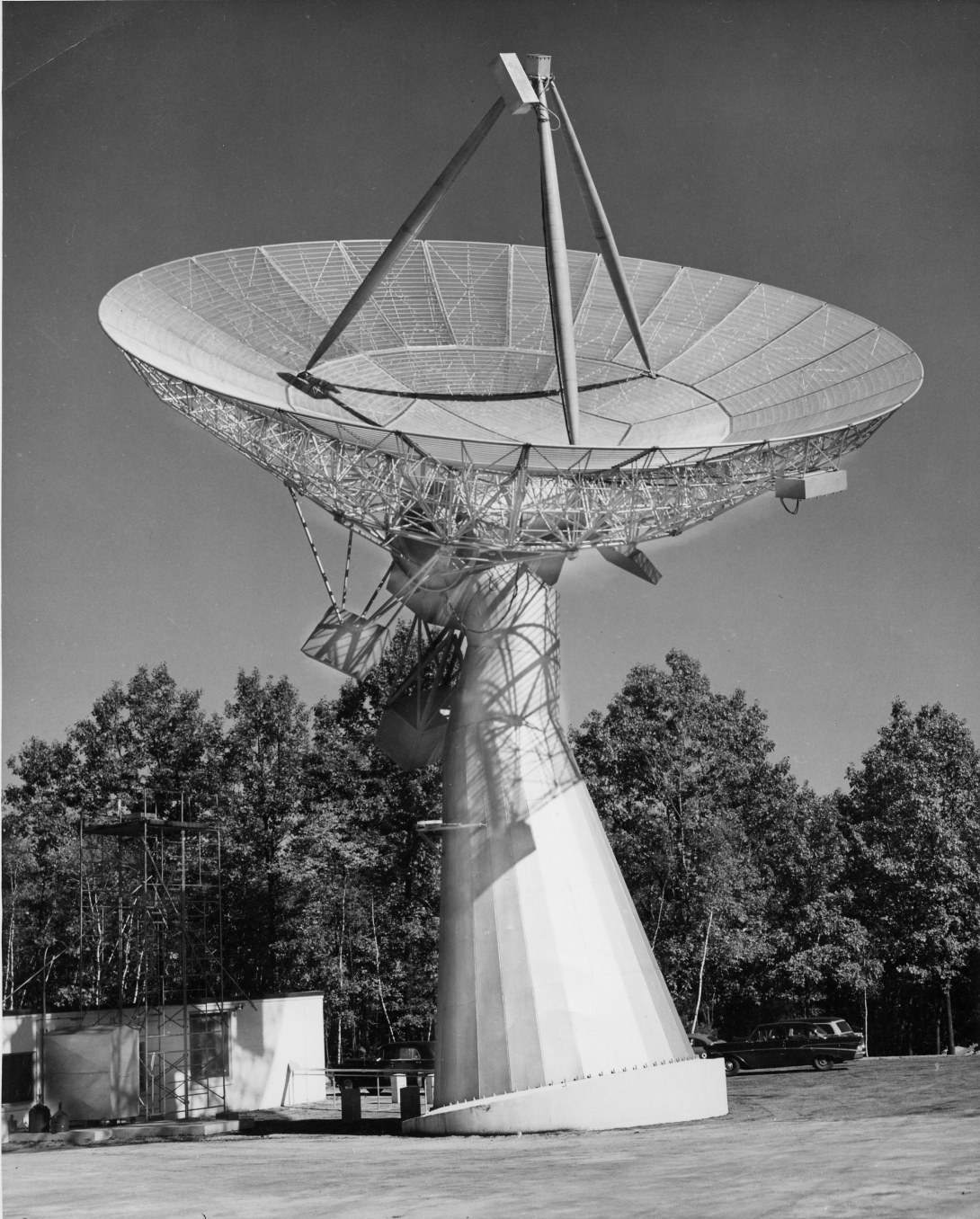 A large dish antenna pointed skyward. The dish is roughly twice the size of a trailer next to its base, and stands above the treetops.