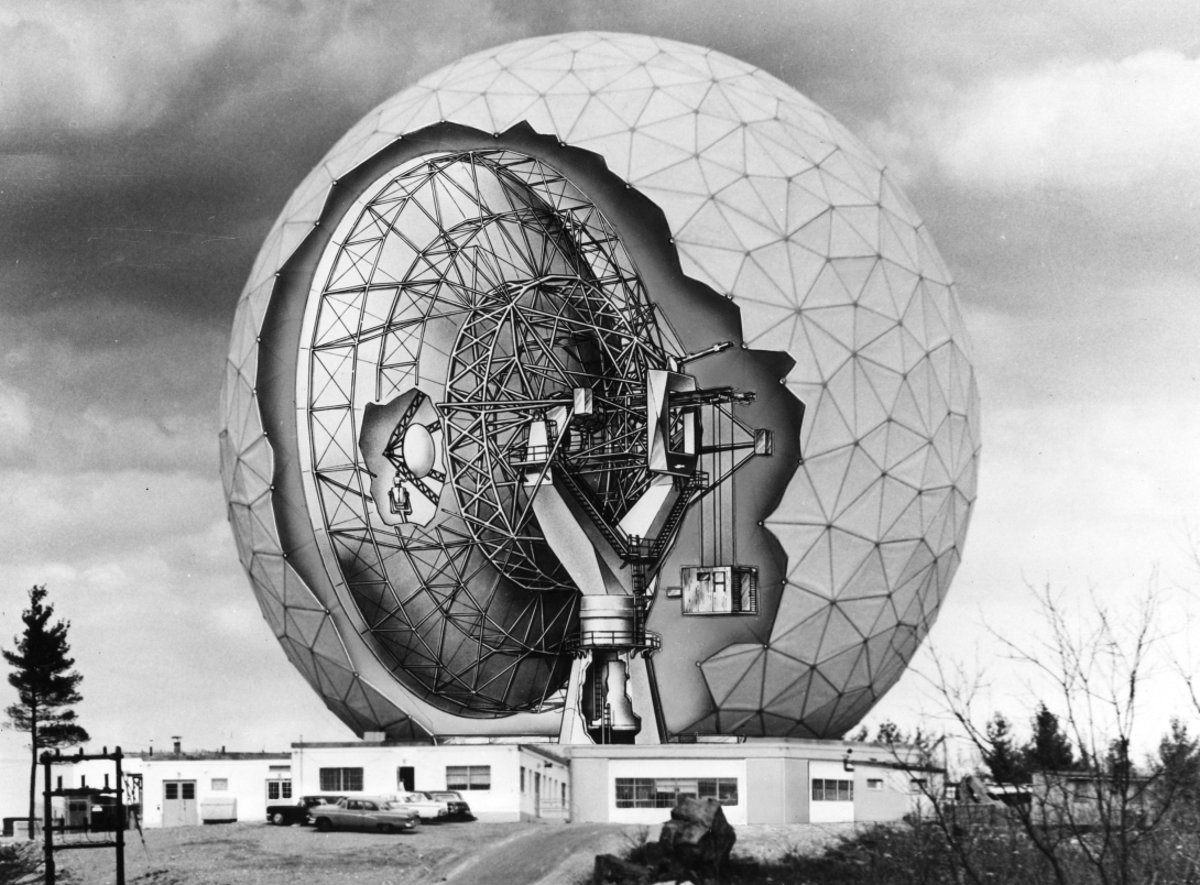 Photograph of a facility where a single story building sits in front of a giant geodesic sphere, roughly 9 stories tall. Overlaid over the sphere is a cutaway drawing which dshows the dish antenna inside.