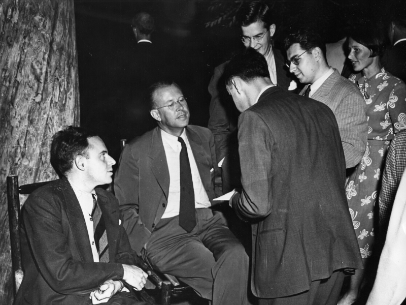 Five well-dressed men and one woman chat during a party. Morrison is one of two men seated at far left; the other stand.